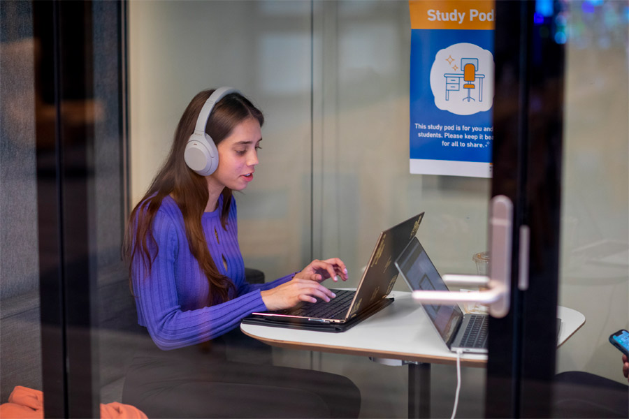 A New York Tech student using headphones sits at a table in a study pod, using a laptop.