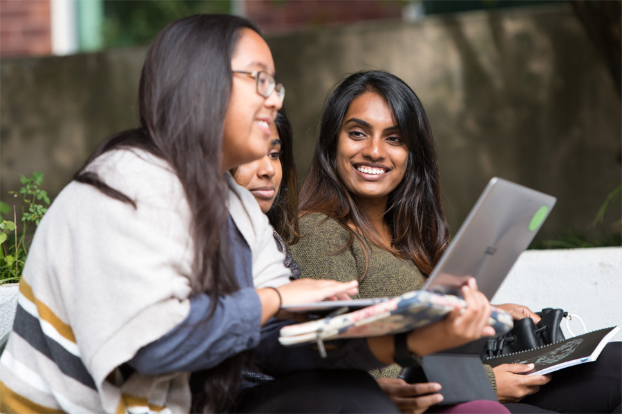 Three smiling New York Tech Honors College students sit down while holding laptops and notebooks.