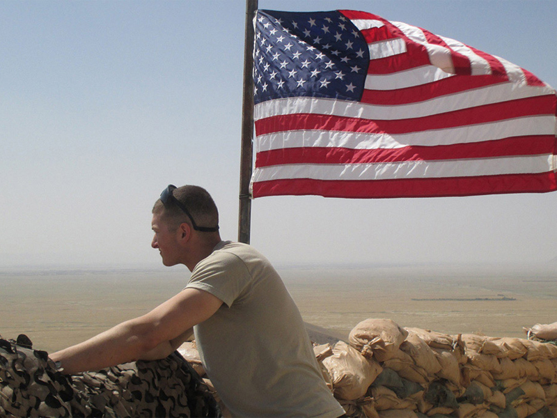 Sergeant Mitchell Stogel standing behind a sandbag wall with an American flag in the background.