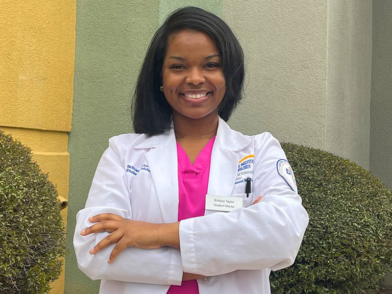 Alum Brittany Taylor stands in a lab coat in front of the NYITCOM-Arkansas building.