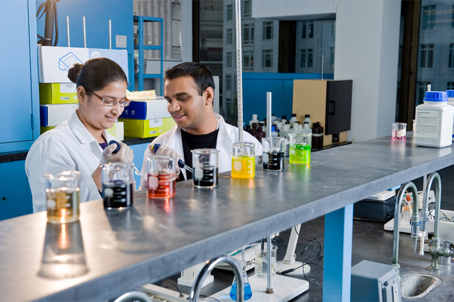 Two students in lab coats working in a lab with colorful chemicals in beakers. 