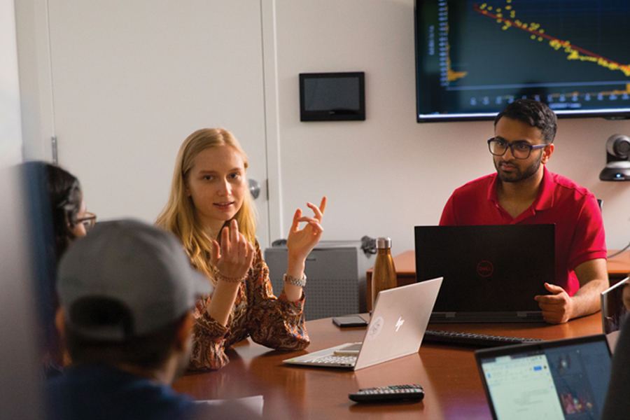 A group of New York Tech School of Management students with laptops discuss sit around a table to discuss concepts while a graph populates in the background.