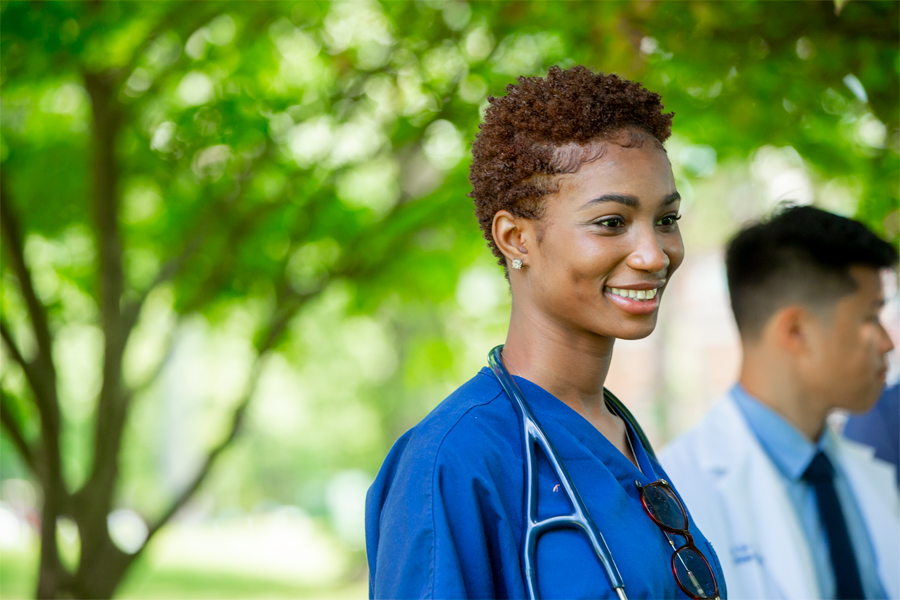 A College of Osteopathic Medicine student wearing a stethoscope smiles while standing outdoors near trees on New York Tech's Long Island Campus.