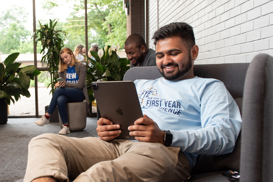 A New York Tech first-year student relaxes in a plush chair in a common area to read something on a tablet.