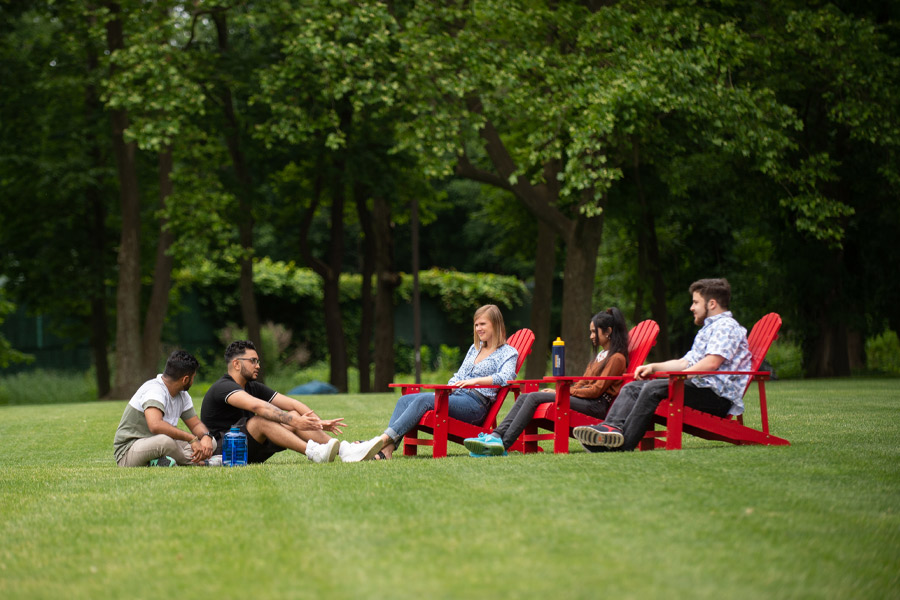 Five New York Tech students relax outdoors on the grass in Adirondack chairs on the Long Island Campus.