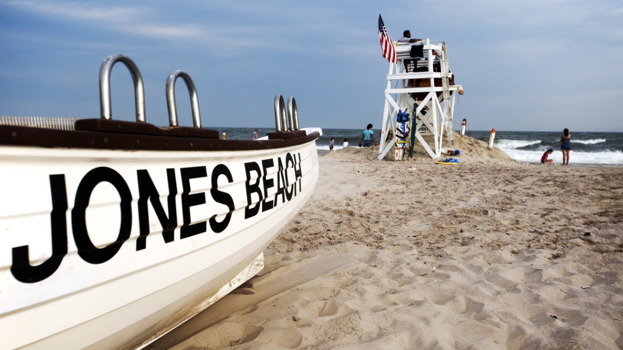 Close up of boat with "Jones Beach" on it with life guard and people in background near ocean.