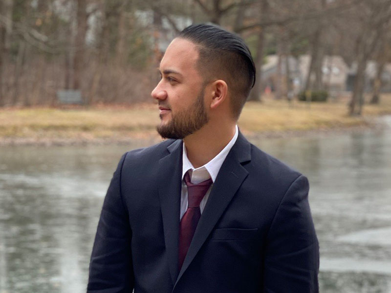 Wearing a suit jacket and tie, New York Tech School of Architecture and Design student Christopher Shagan stands in front of a pond.