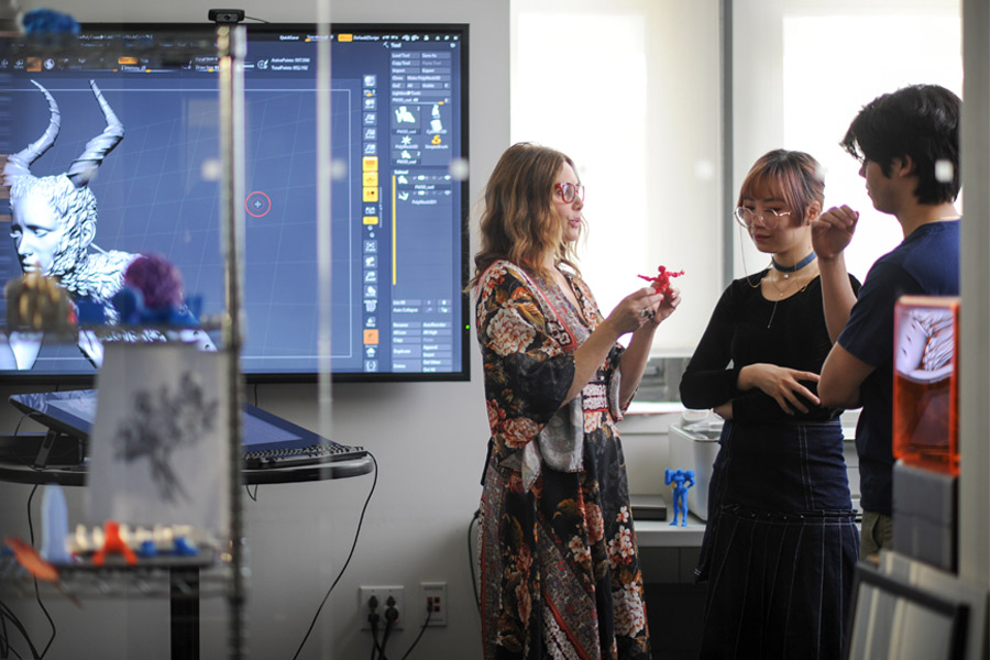 A professor standing in front of a screen with a 3-D graphic converses with two students in one of New York Tech's digital art and design studios.