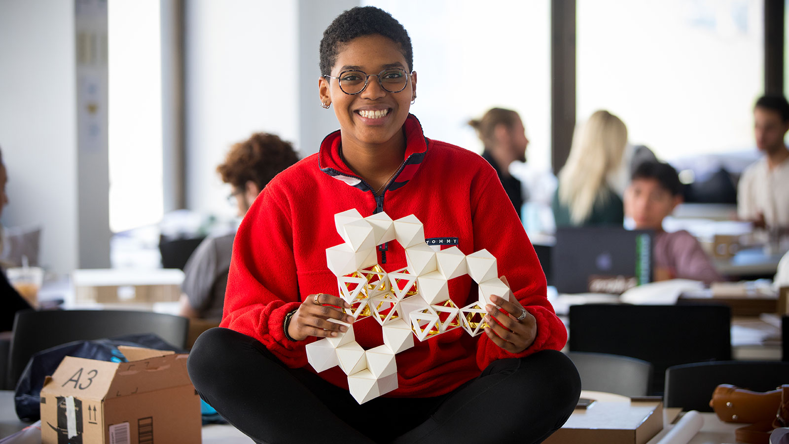 A New York Tech School of Architecture and Design student sits cross legged while holding a 3-D model of a cubed structure.