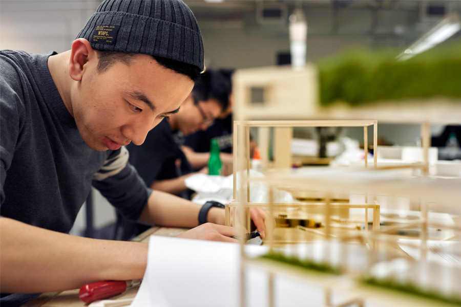 Sitting at a table, a New York Tech School of Architecture and Design undergraduate student focuses on building a 3-D model out of wood pieces.