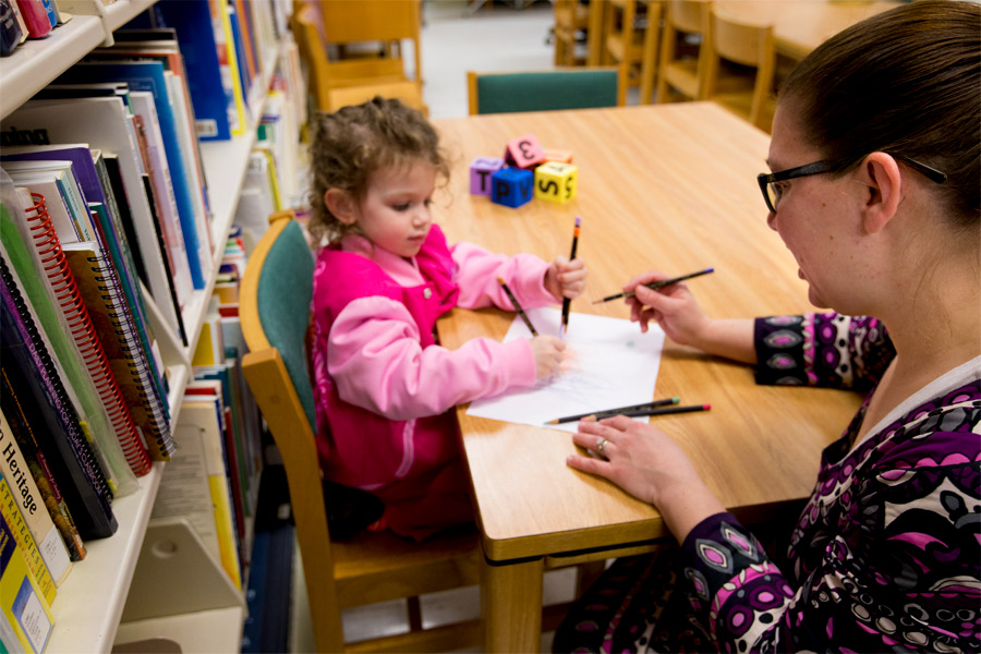 During a field assignment, a New York Tech education student sits at a table in a library to draw a picture with an elementary school-age child.