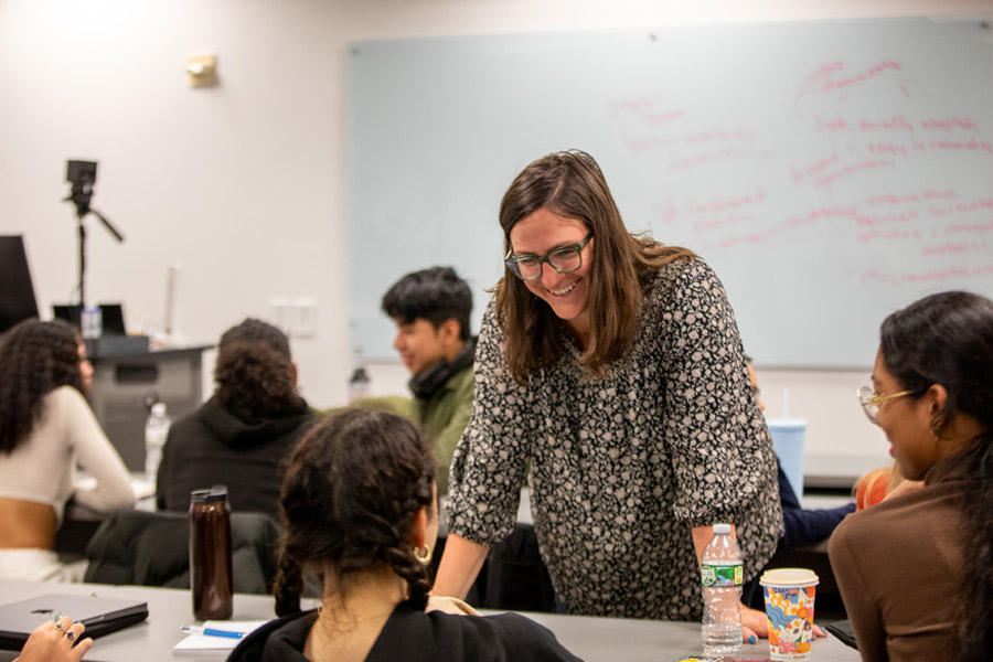 In a classroom for one of New York Tech's College of Arts and Sciences undergraduate programs, a faculty member smiles while conversing with a group of students sitting along a lecture table.