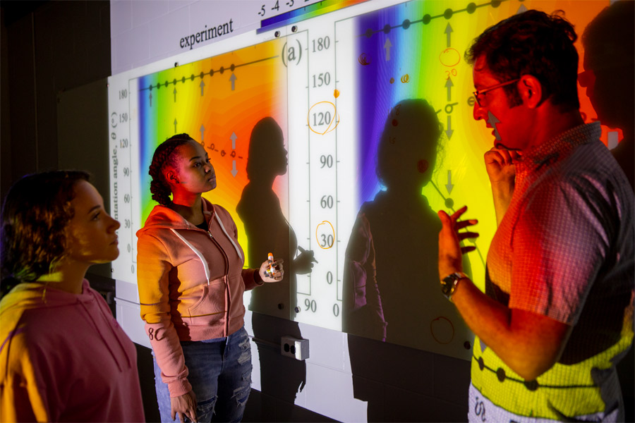 Standing in front of a large projected screen, a faculty member instructs two students in using an applied mathematics software program.