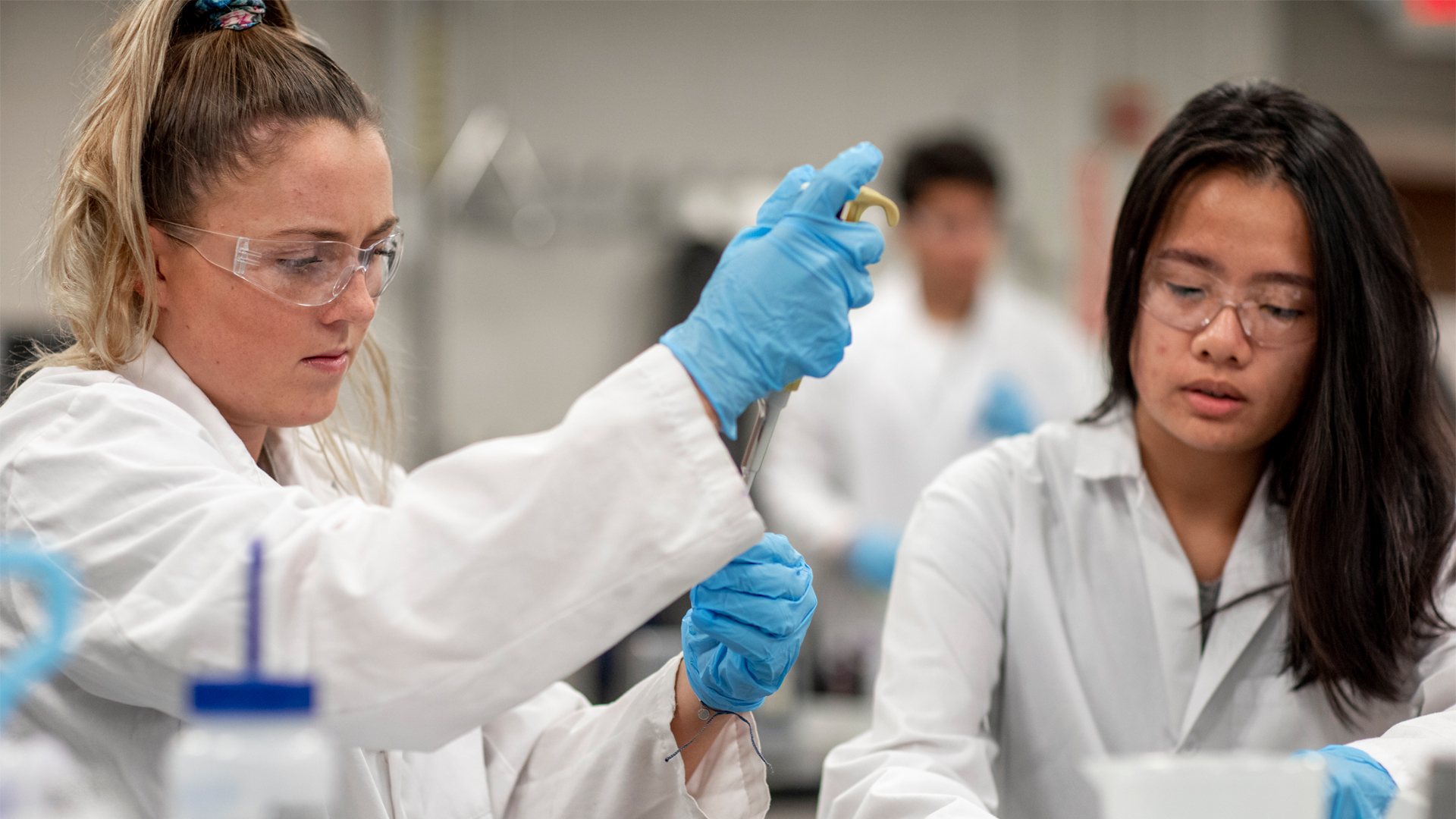 In one of New York Tech's science labs, two College of Arts and Sciences students wearing lab coats, gloves, and safety glasses get experience using the latest research equipment.