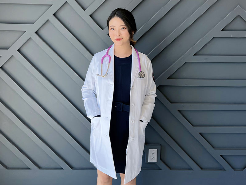 New York Tech College of Arts and Sciences student Selin Park stands against a geometrically patterned wall while sporting a lab coat and stethoscope.