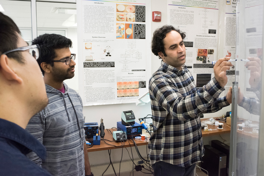 A faculty member and two students work together in a lab with poster boards of projects in the background.