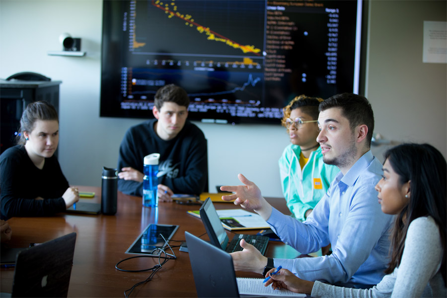 People in a business conference room with a large table with a graph on a large screen in the background.