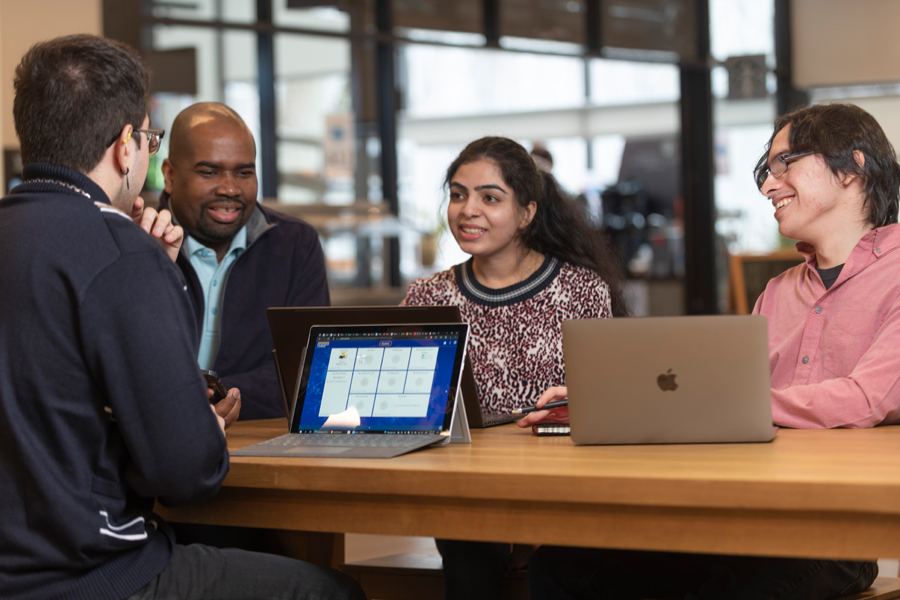 Four students working in a lounge at a table with laptops.