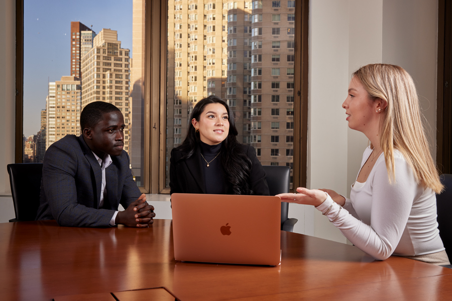 Three students in business attire sitting and talking in a conference room at a large table on the New York Tech NYC campus.