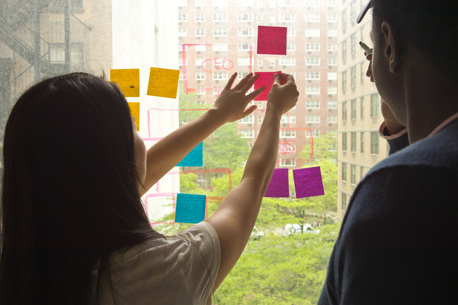 Two student using a window to create a flow chart with dry erase markers and post-its. 