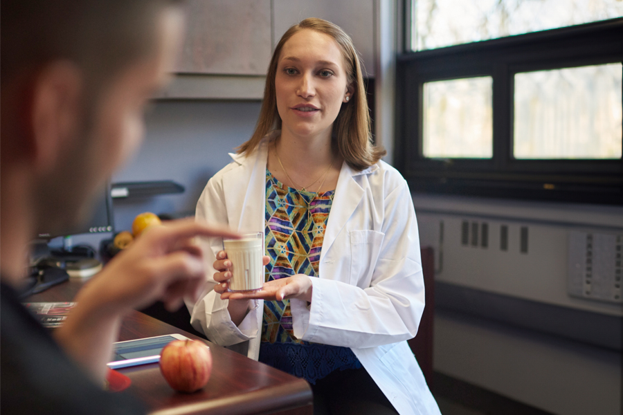 Two students in a nutrition lab. one of holding a glass of milk and there is an apple on the table. 