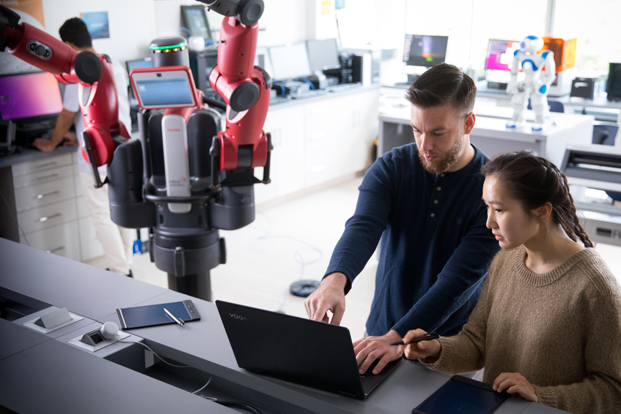 Two student work on a laptop in a robotics lab. There is a black and red robot in the background.