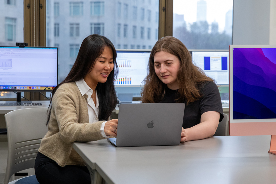 Two students working together in a computer lab sitting at a table with a laptop. 