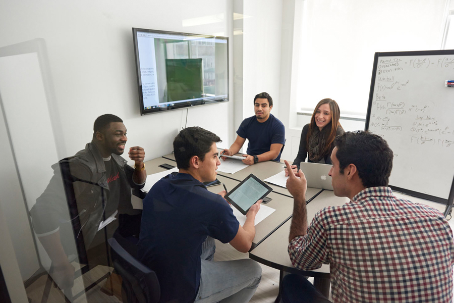 A group of 5 students seated in a conference room with whiteboard in the background. 