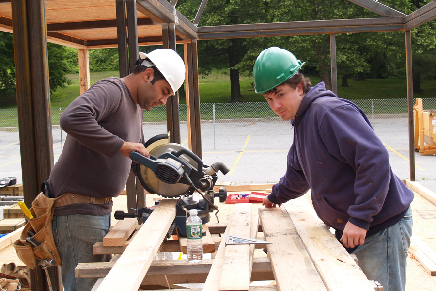 Two people in hard hards cutting wood planks outside.