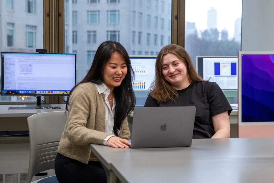 Two students in a computer lab, sitting at a table in front of a laptop. 