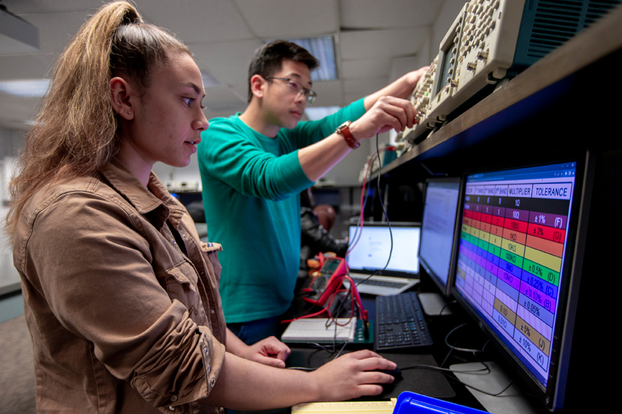 Two students in a lab working on electrical equipment and monitoring on a computer. 