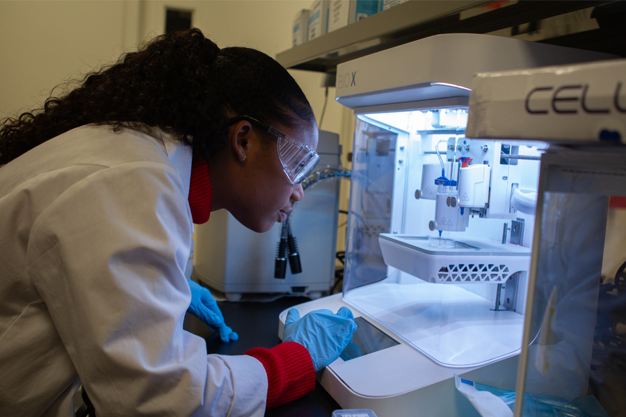 In New York Tech's Bio-Nanotechnology and Biomaterials Lab, a female student wearing a lab coat, safety glasses, and latex gloves observes a 3-D printer creating a prototype for a project focusing on bone healing.