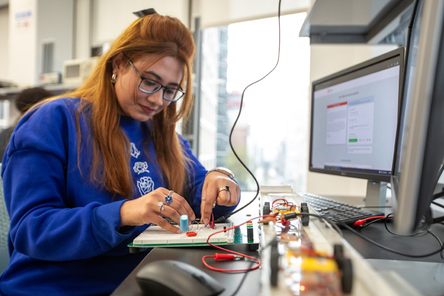 A female computer science student at New York Tech works on a breadboard in front of a computer.