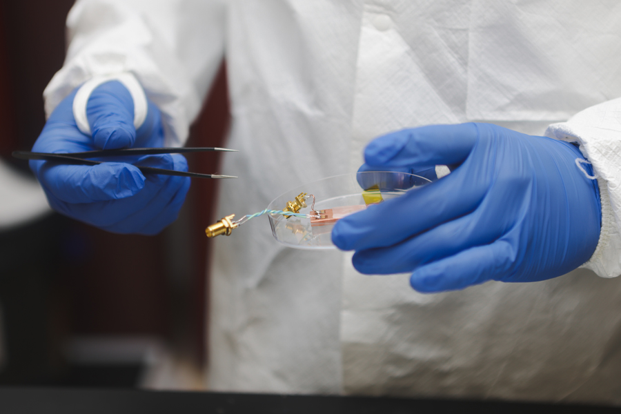 A student wearing blue latex gloves and a cleanroom suit handles electrical components with forceps and a petri dish in a research lab in New York Tech's College of Engineering and Computing Sciences.