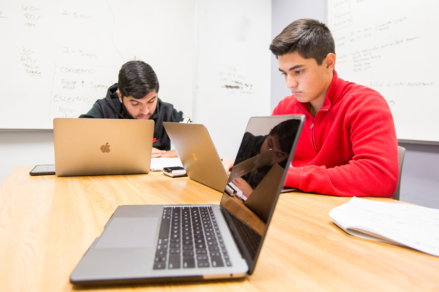 Two students in a study lounge sitting at a large table with two whiteboards in the background.