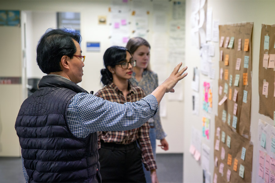 A faculty member explains how return on investment works to two New York Tech students, using a series of sticky notes and kraft paper.