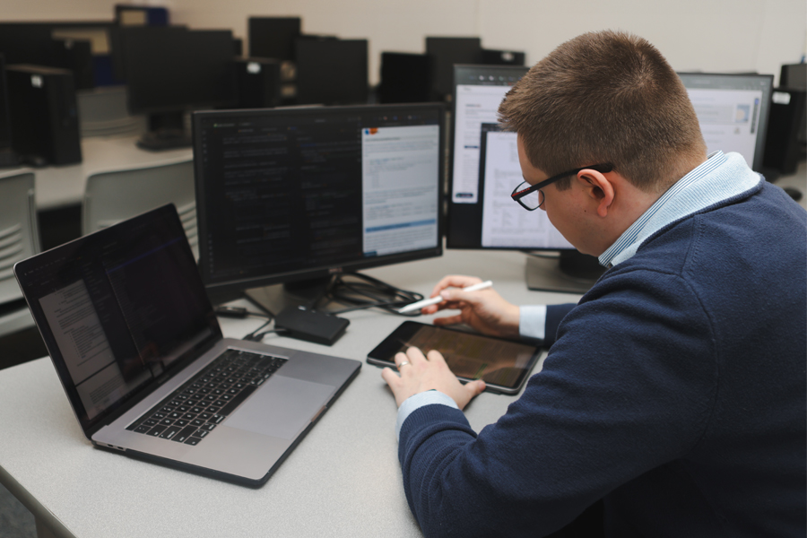 Man at a desk with laptop and computer screens looking at an iPad