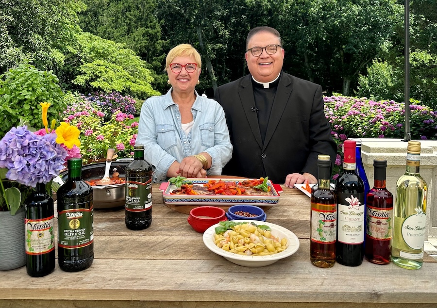 Two people standing behind platters of food and oil and sauce bottles. There is a garden with purple flowers in the background.