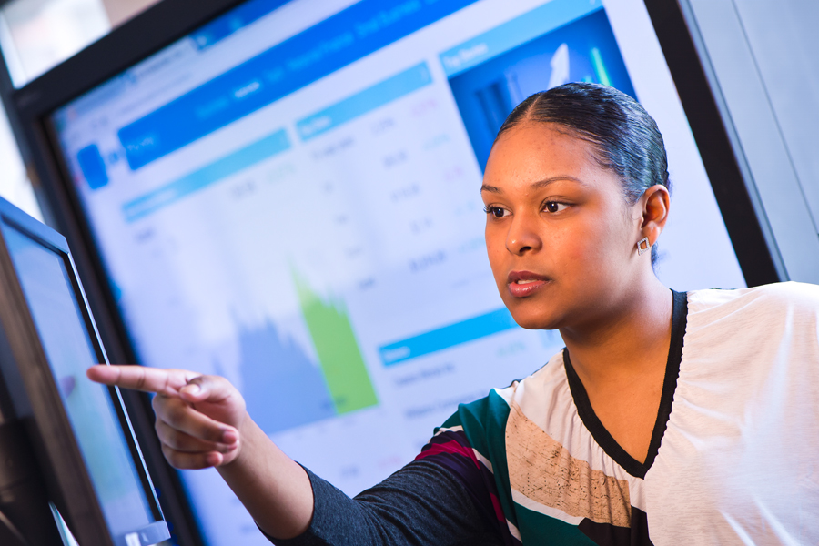 A New York Tech School of Management student points to a computer screen while a larger screen behind displays a dashboard with graphs.