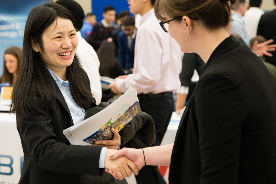 A New York Tech School of Management student wearing a blazer and holding a brochure shakes hands with a recruiter during a job fair.