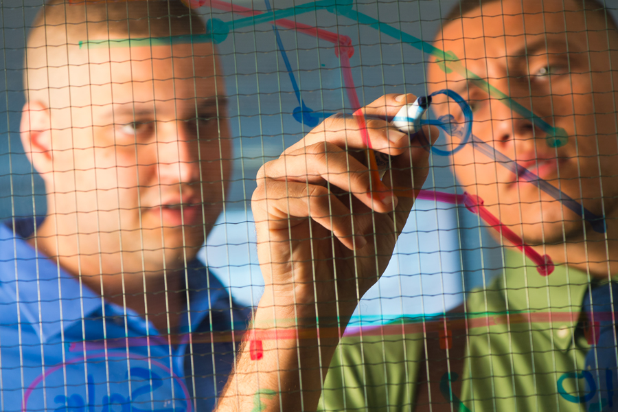 Two New York Tech School of Management students in collared shirts draw a graph with colored markers on a clear plastic surface.