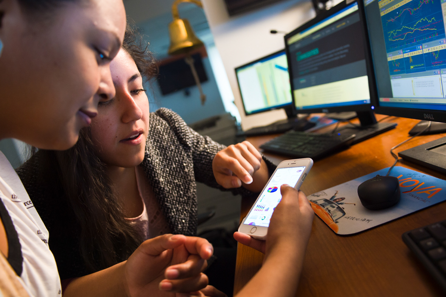 Two student working in a computer lab looking at a smartphone with computer screens filled with graphs and charts in the background. 