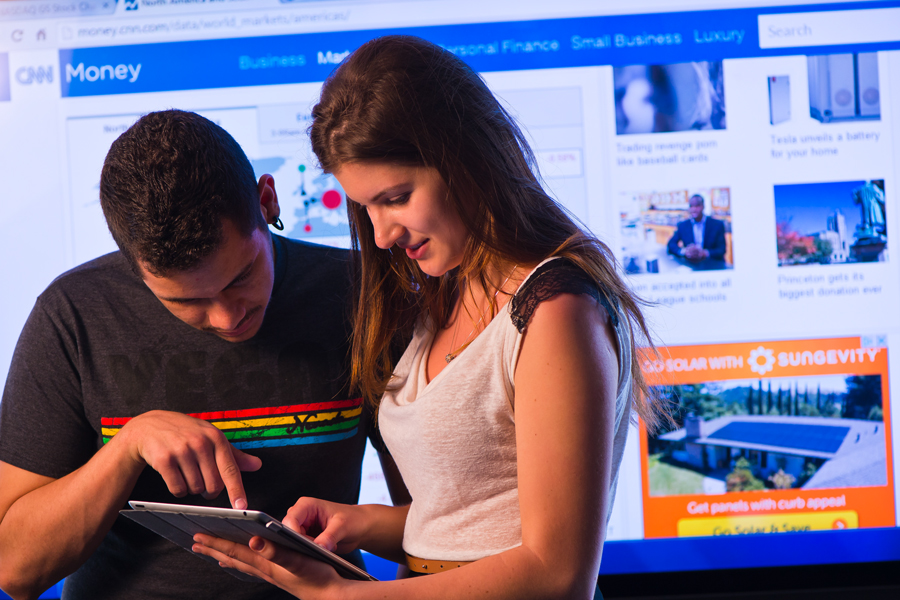 Two students working on a tablet with a large screen behind them.