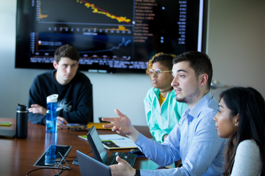 People in a business conference room with a large table with a graph on a large screen in the background.