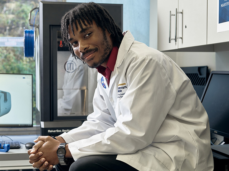 New York Tech Mechanical Engineering, B.S. alum Lincoln Dover sits in a computer lab while wearing a lab coat.