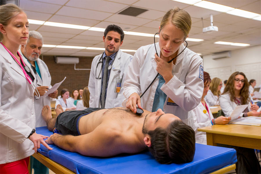 Four students in white coats practicing on a patient laying down on a medical examination table in a classroom environment.