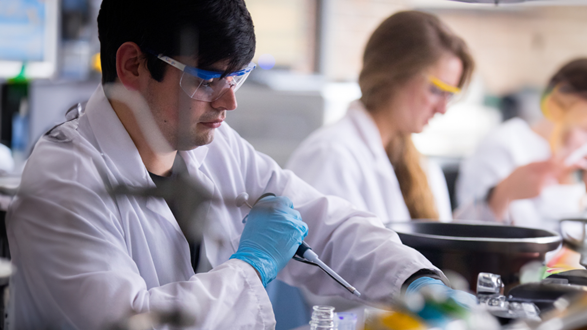 Man in a lab coat and safety gear using a tool in a chemistry lab. People are blurred in background behind him.