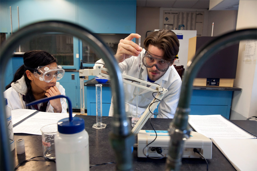 Two students in a lab wearing safety gear, one holding a dropper over a testing cylinder.