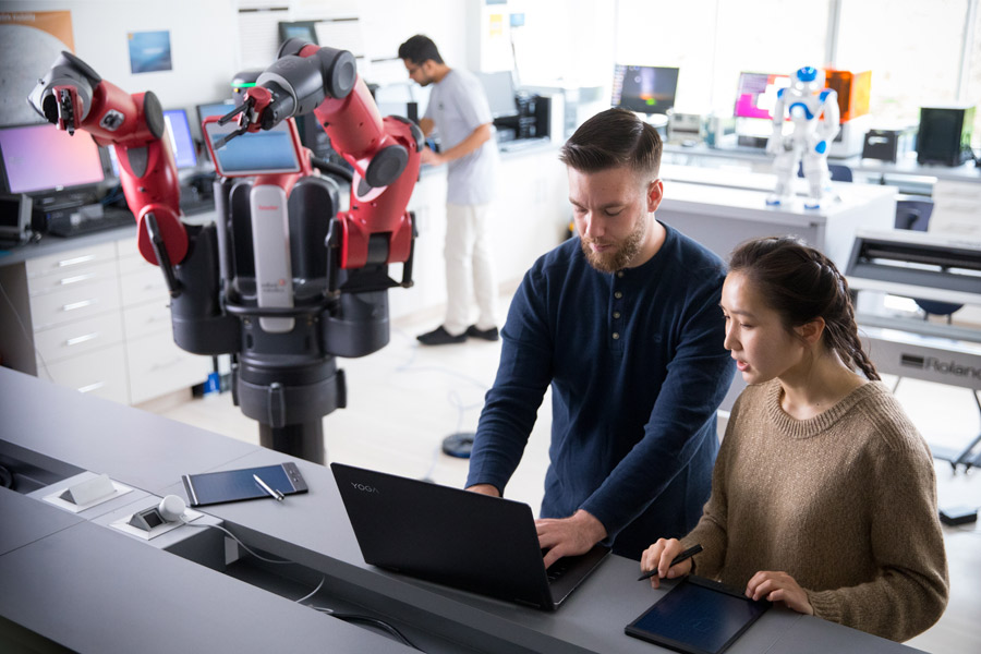 Students in a robotics lap, one in the back and two in the front on a laptop. They are next to a large red robot with two arms.