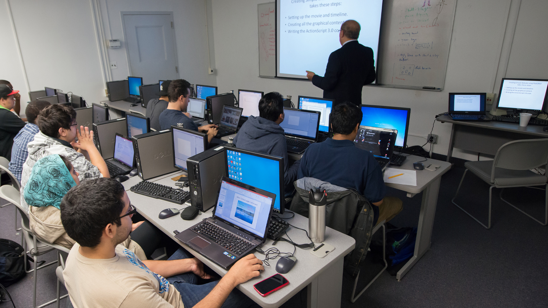 Students work in a computer classroom while a professor teaches a course.